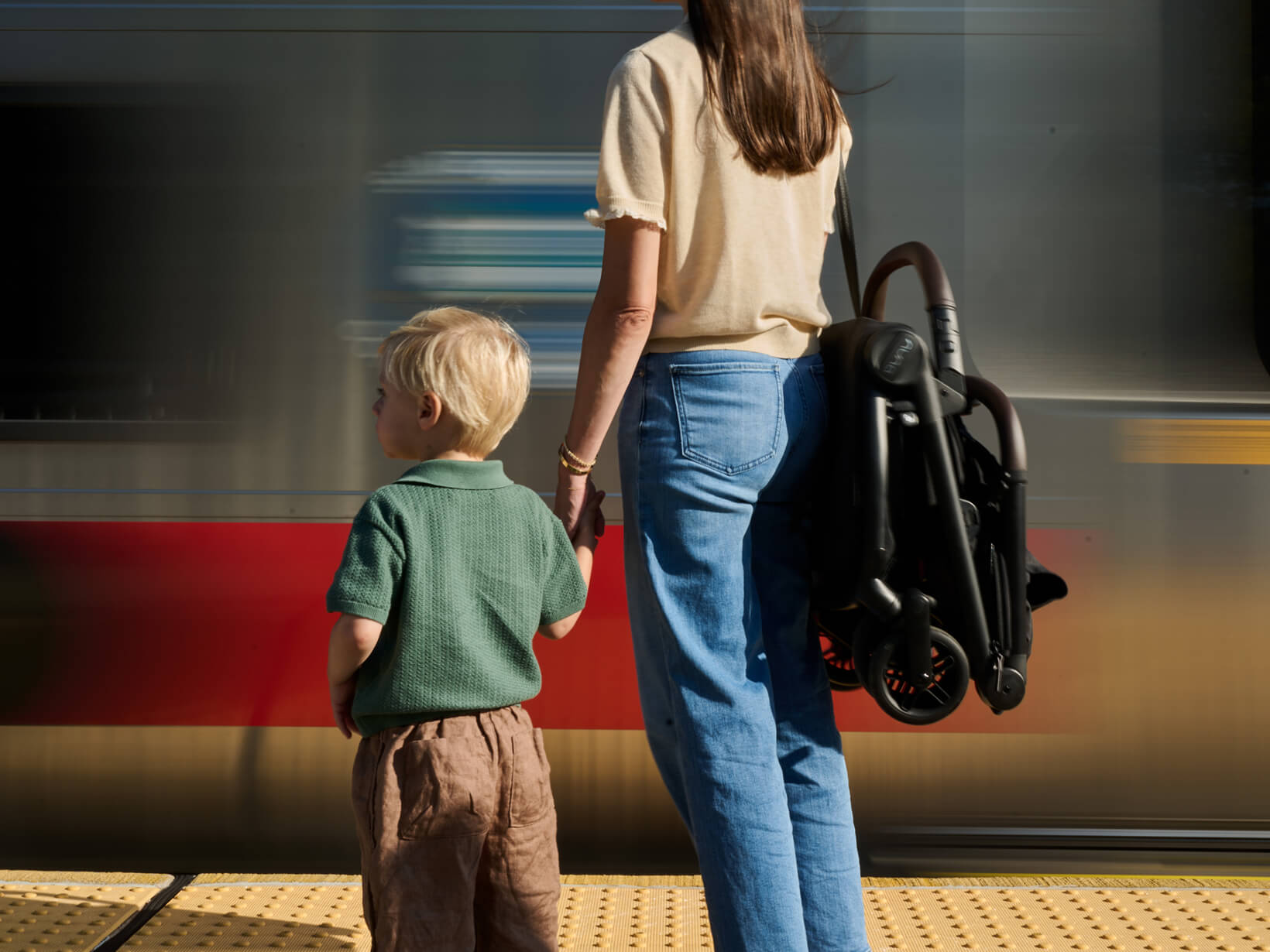 A mother holds her toddlers hand at a train station while wearing the folded VIAA cabn puschair over her shoulder.