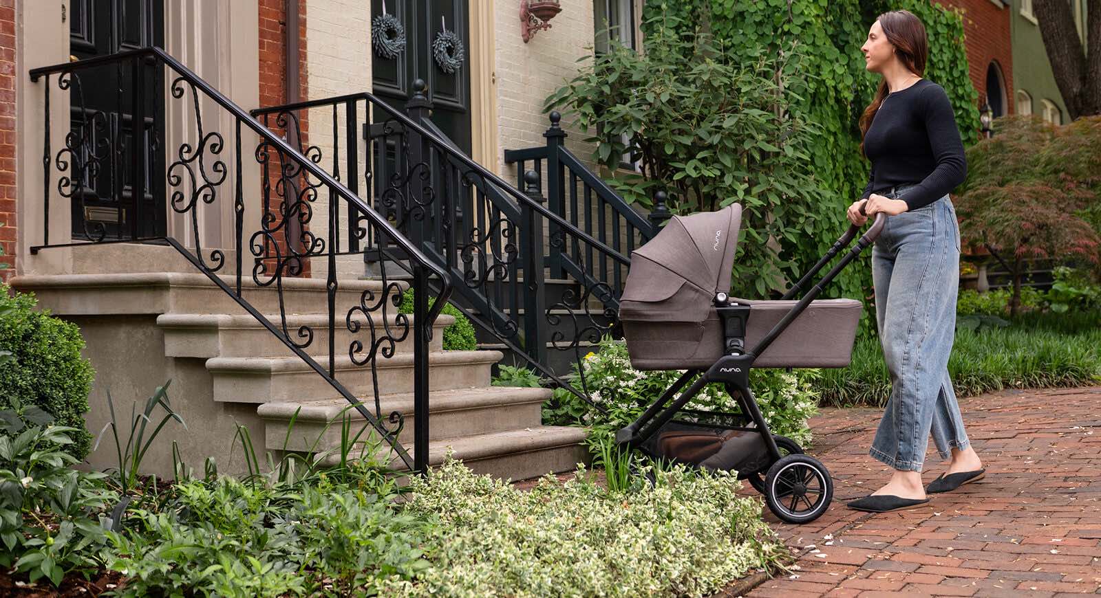 Woman pushing the Nuna Mixx Next stroller with carry cot attachment down a tree-lined city sidewalk.