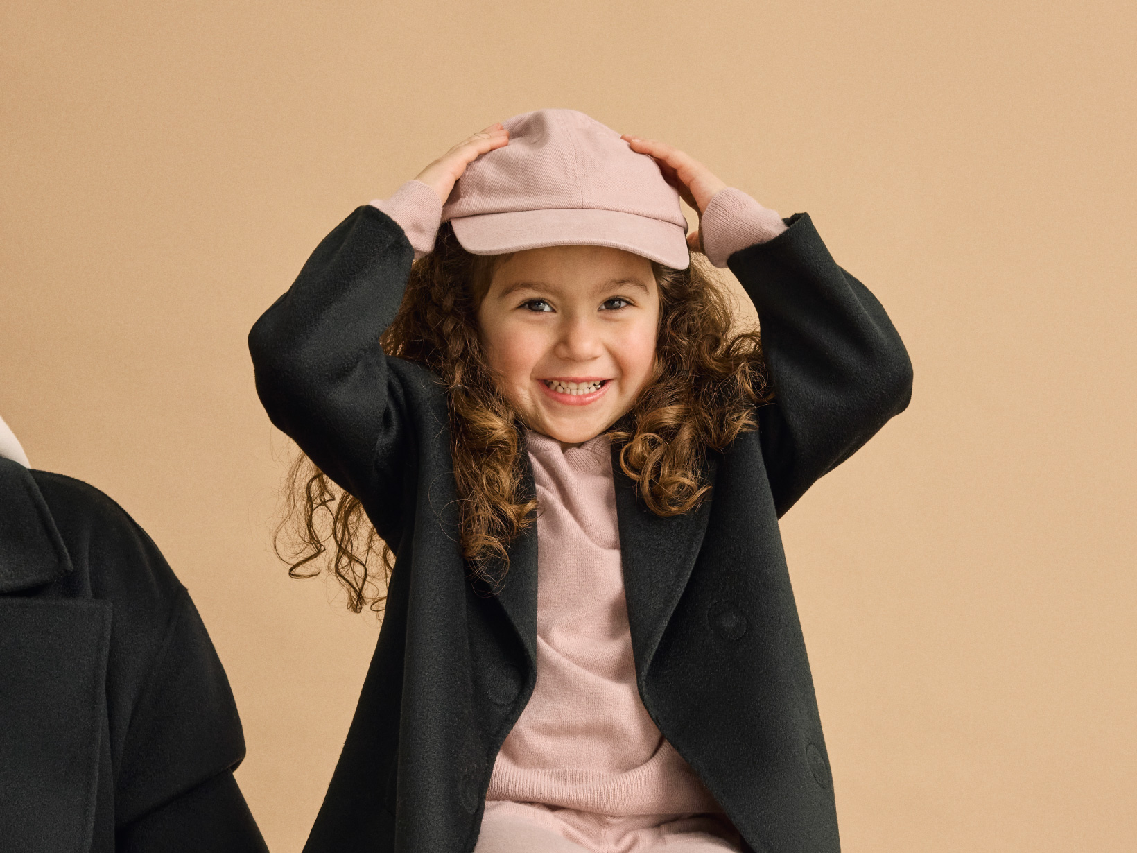 A young girl smiling with her hands on her head wearing a Nuna Cashmere Coat in Shadow fashion and Nuna Child's Two-Piece Sweater Set in Dusty Mauve