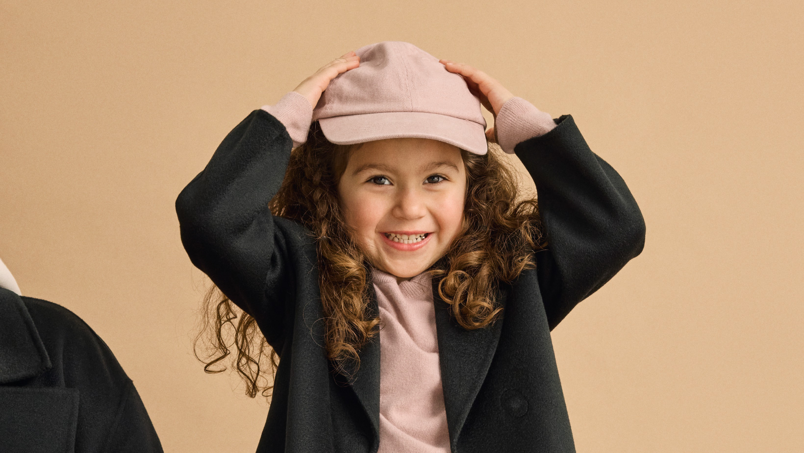 A young girl smiling with her hands on her head wearing a Nuna Cashmere Coat in Shadow fashion and Nuna Child's Two-Piece Sweater Set in Dusty Mauve