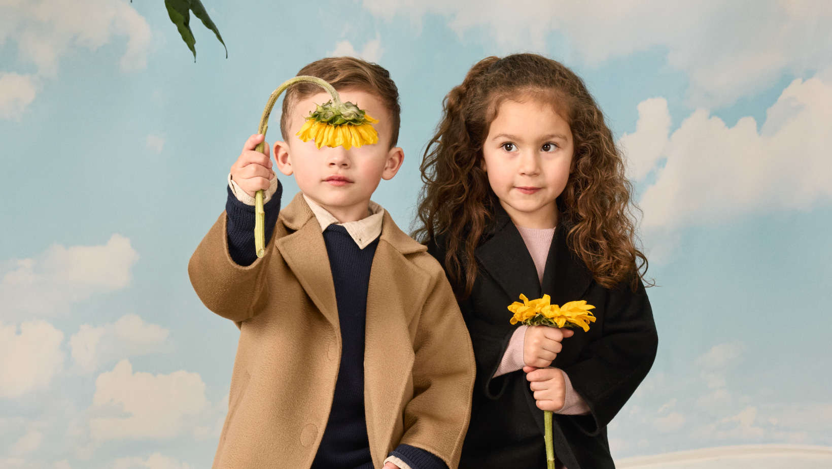 A girl and boy holding sunflowers wearing matching Nuna Cashmere Coats, in Camel and Shadow fashion