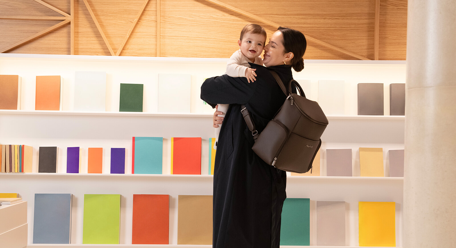 A mother holds her young child in a bookstore while wearing the Nuna Essentials Bag. 