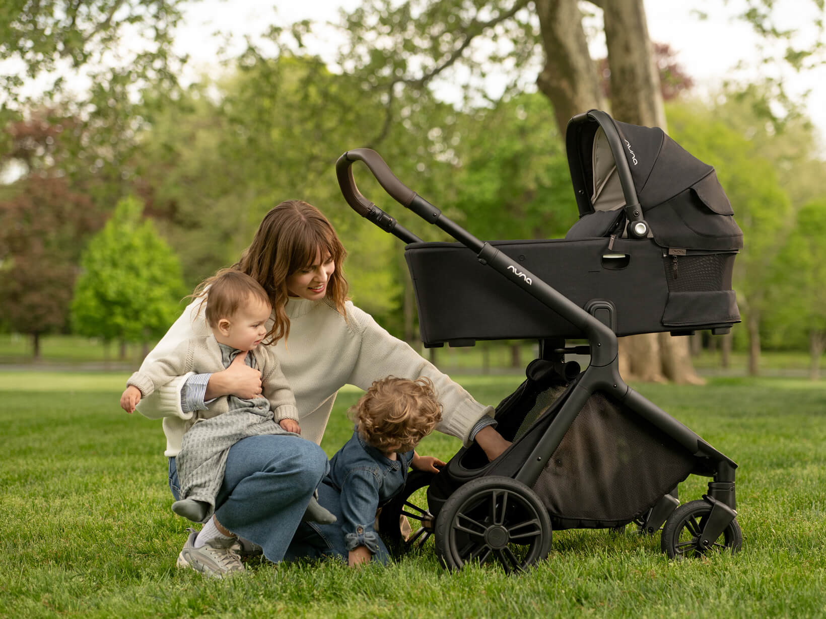 A woman plays at the park with her two young kids. The Demi icon pushchair with carry cot attachment sits nearby.