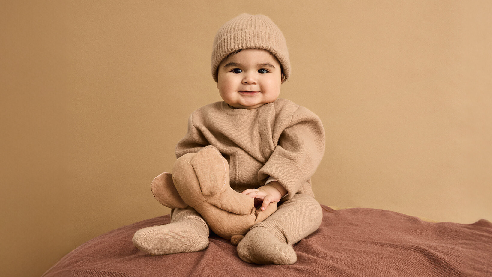 A baby with a teddy wearing the Nuna Cashmere Asym Sweater, Knit Cap and Socks in Tan sitting on a Cashmere Blanket in Sienna