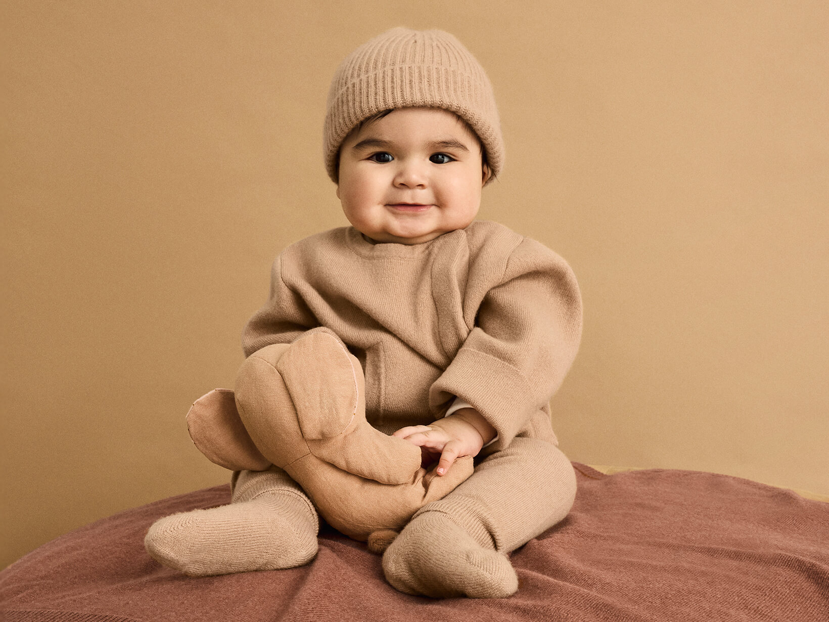 A baby with a teddy wearing the Nuna Cashmere Asym Sweater, Knit Cap and Socks in Tan sitting on a Cashmere Blanket in Sienna