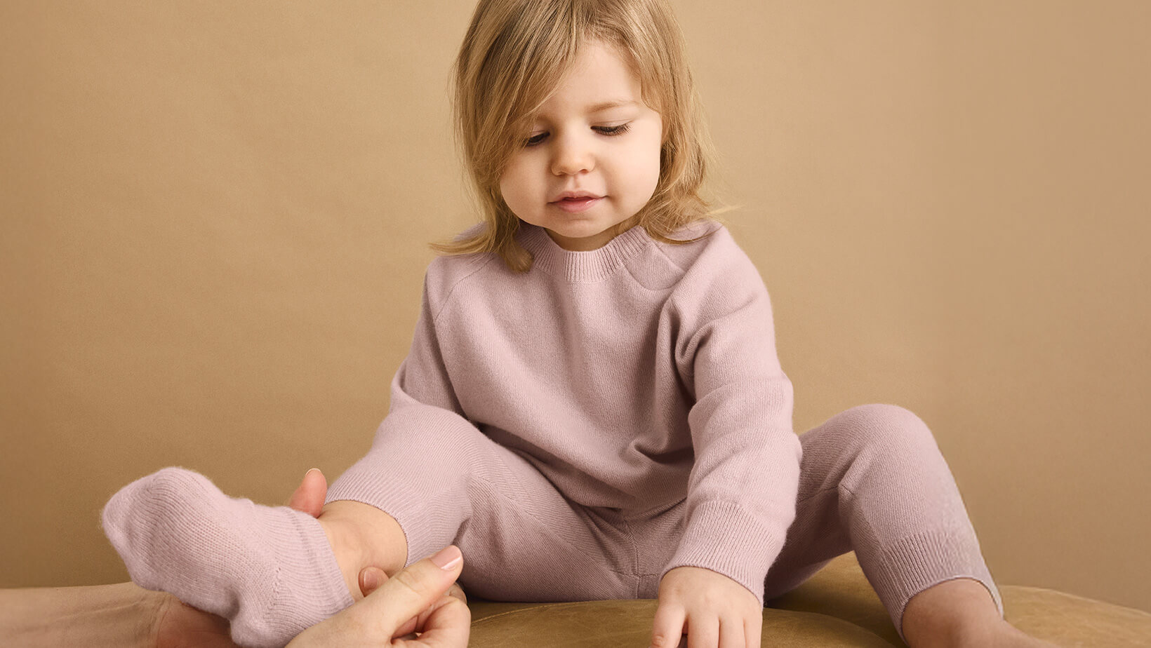 A young girl sat on a cushion with a woman putting a sock on her wearing Nuna Cashmere Two-Piece Set and Socks in Dusty Mauve