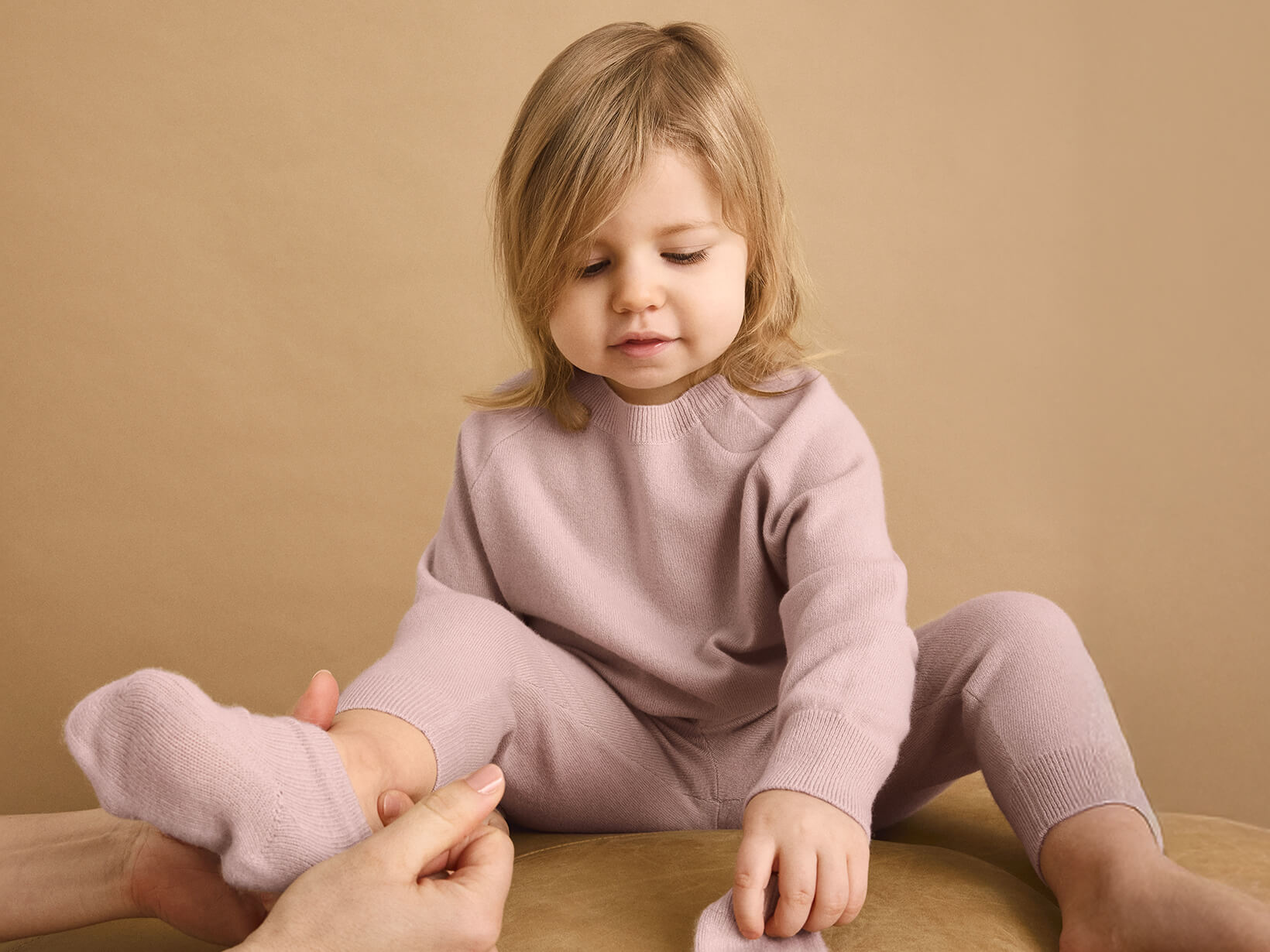 A young girl sat on a cushion with a woman putting a sock on her wearing Nuna Cashmere Two-Piece Set and Socks in Dusty Mauve