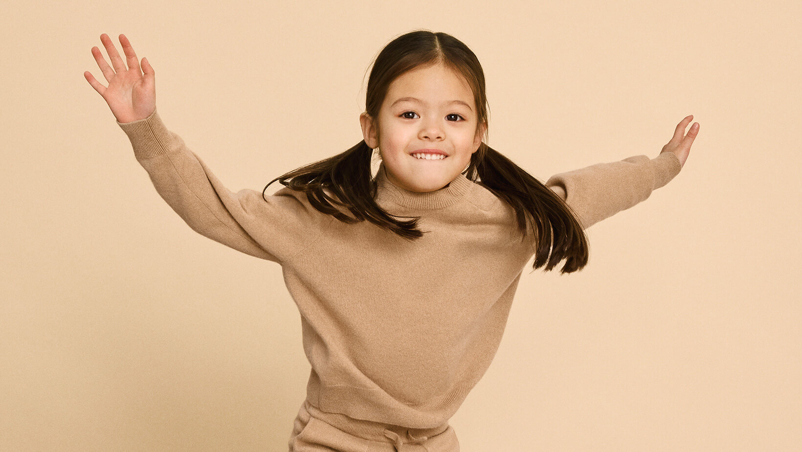 A young girl jumping on a couch wearing Nuna Cashmere Two-Piece Sweater Set in Tan