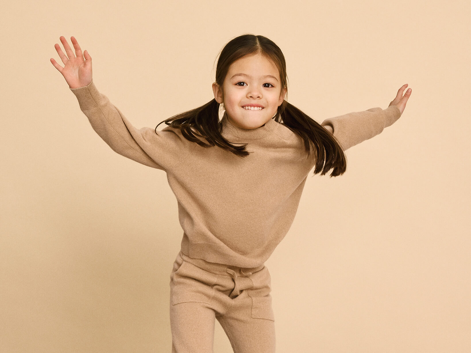 A young girl jumping on a couch wearing Nuna Cashmere Two-Piece Sweater Set in Tan
