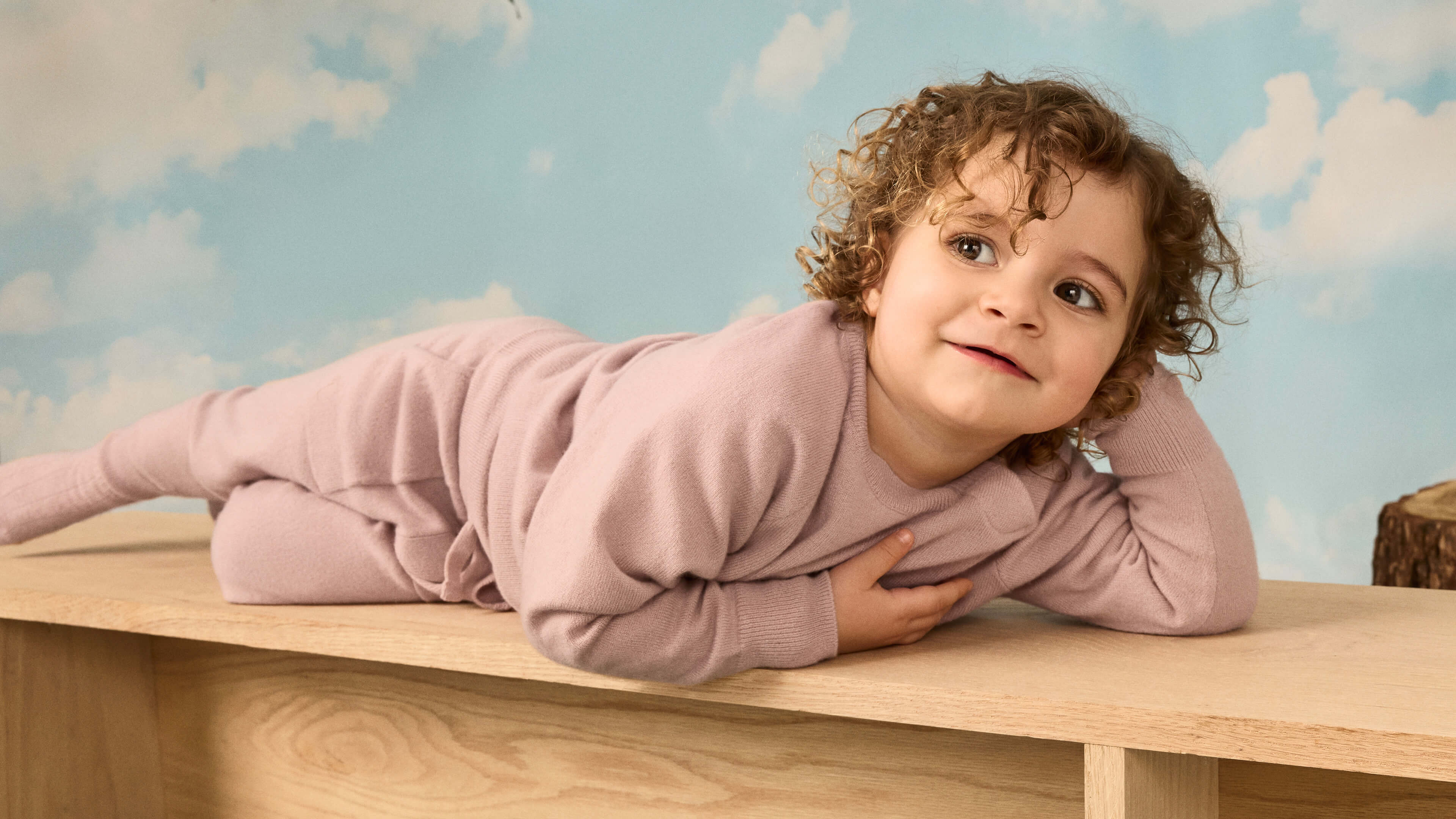 A young girl lying on a wooden bench wearing the Nuna Cashmere Two-Piece Sweater Set and Socks in Dusty Mauve