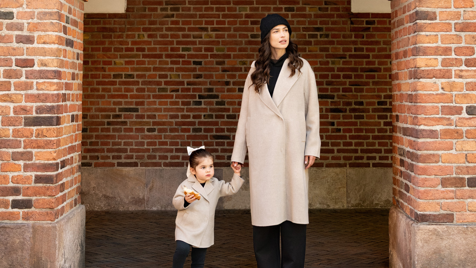 A woman and a young girl standing in a brick archway holding hands wearing matching Nuna Cashmere Coats in Stone fashion