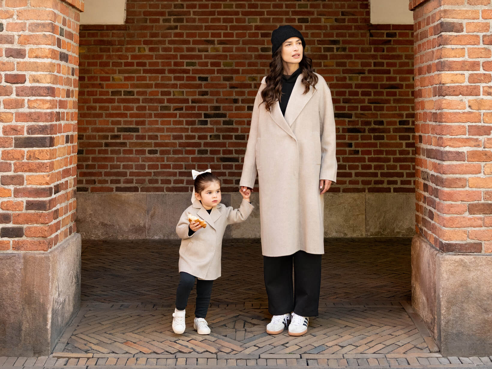 A woman and a young girl standing in a brick archway holding hands wearing matching Nuna Cashmere Coats in Stone fashion