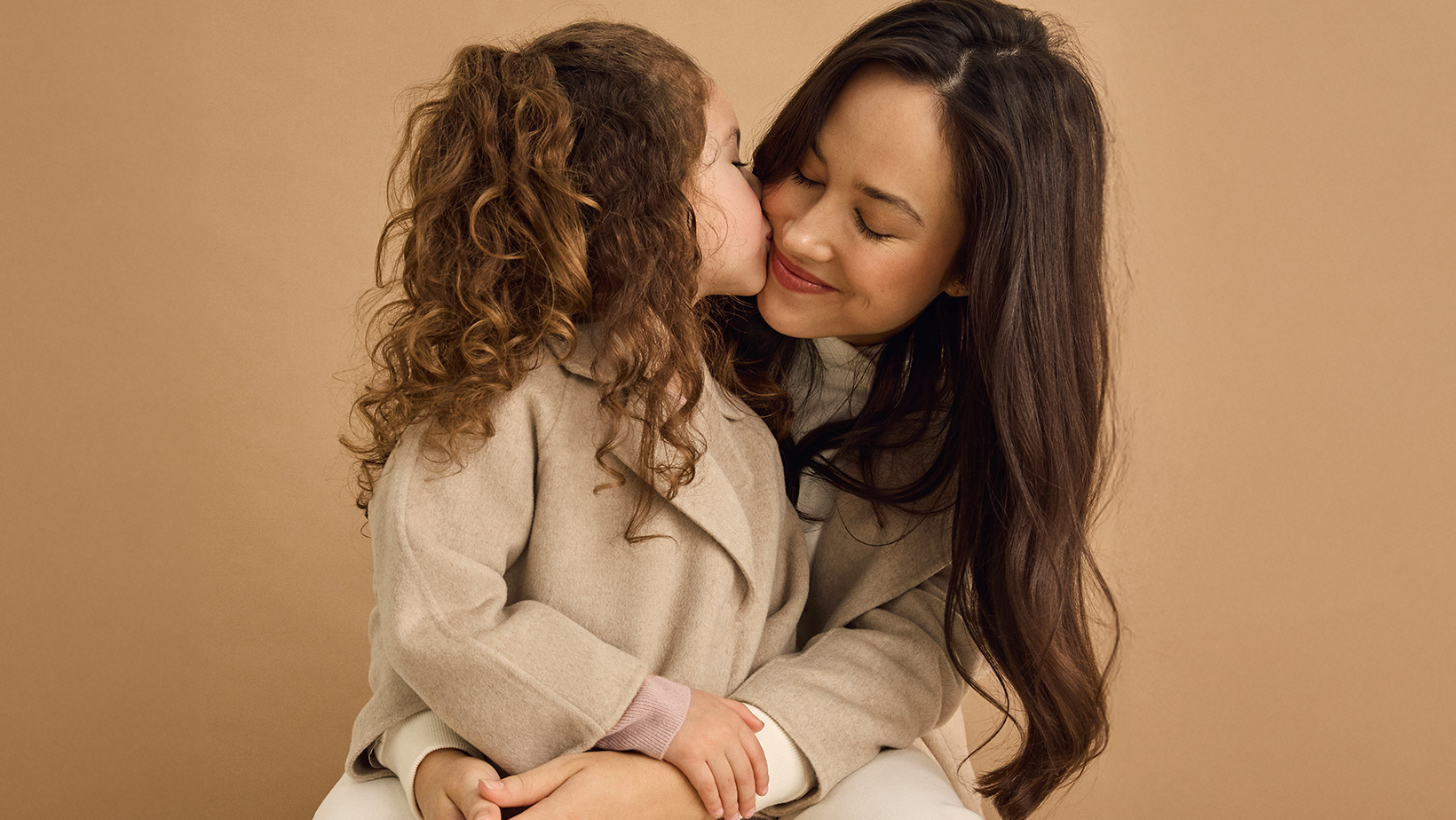 A young girl standing in front of a woman giving her a kiss and wearing matching Nuna Cashmere Coats in Stone fashion