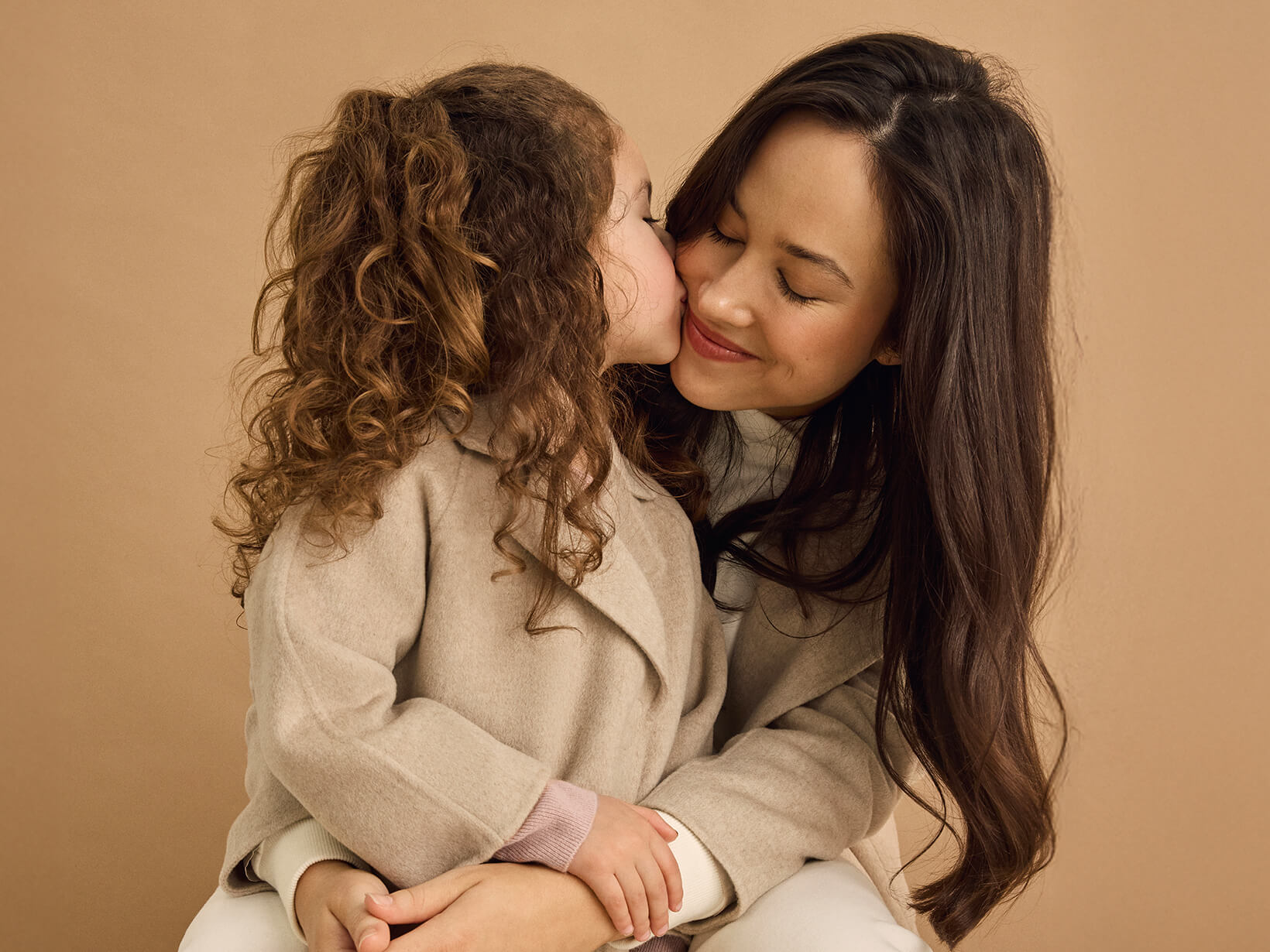 A young girl standing in front of a woman giving her a kiss and wearing matching Nuna Cashmere Coats in Stone fashion