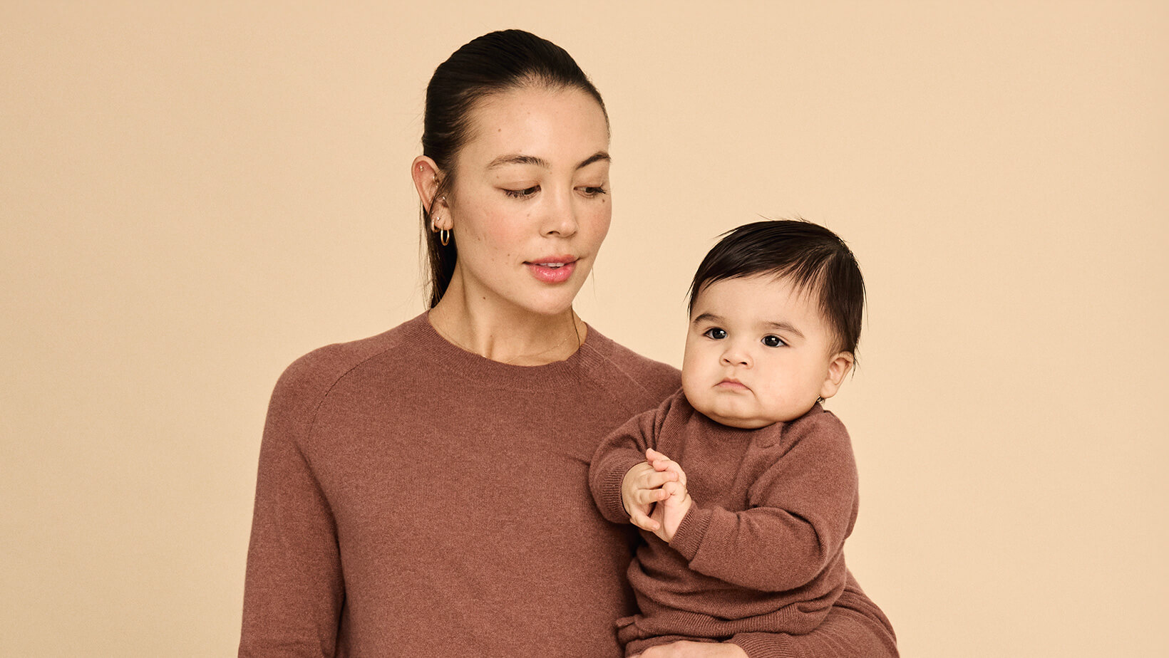 A woman standing in front of a couch holding a baby wearing matching Nuna Cashmere Two-Piece Sweater Sets in Sienna