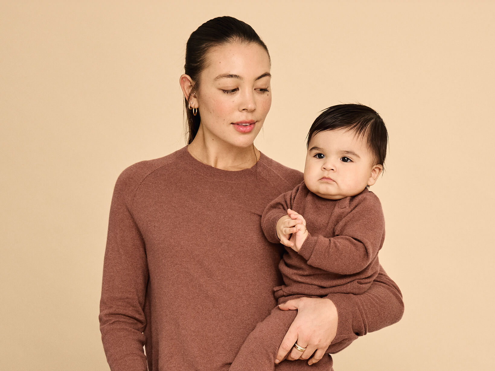 A woman standing in front of a couch holding a baby wearing matching Nuna Cashmere Two-Piece Sweater Sets in Sienna