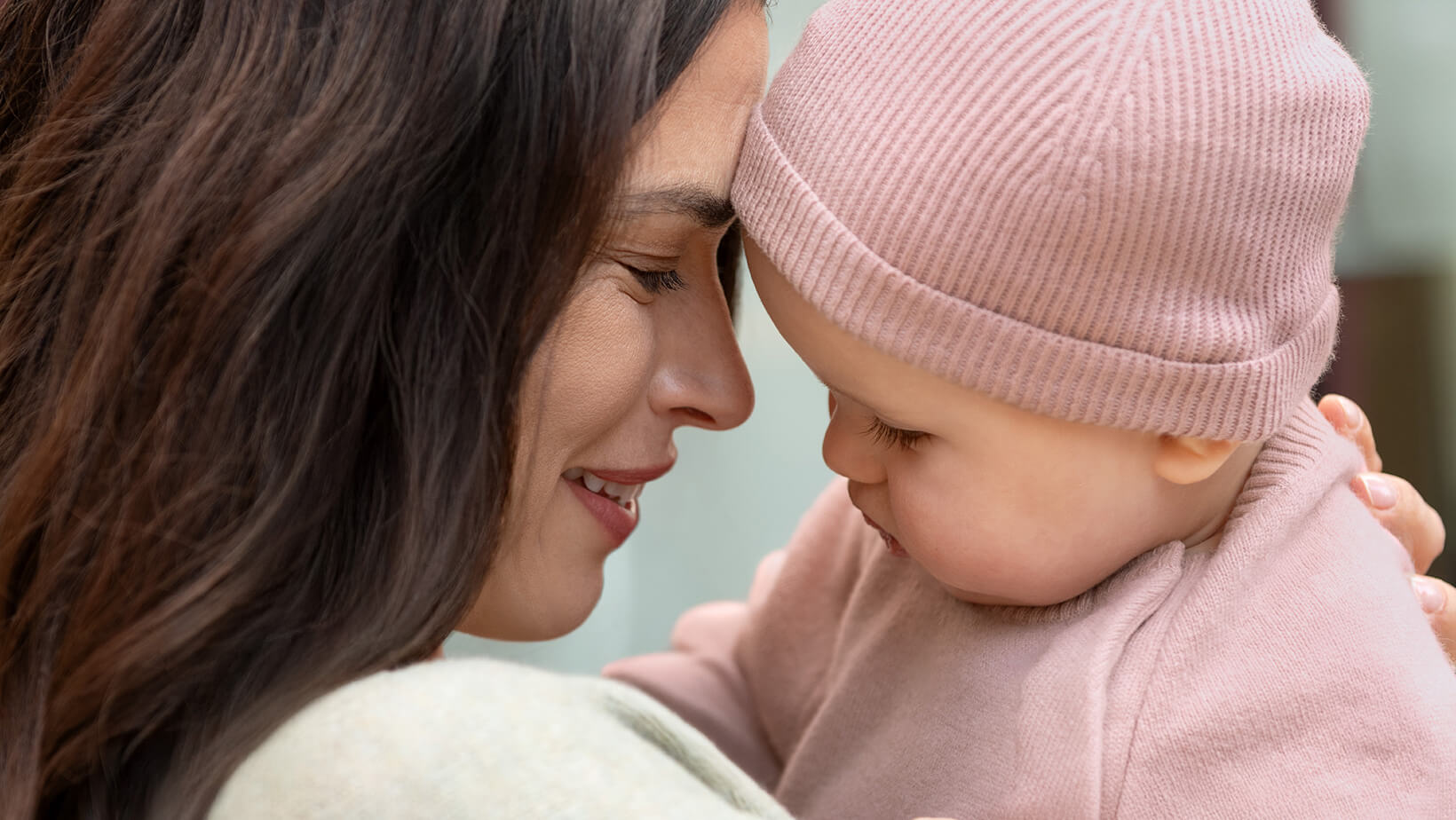 A close up of a woman holding a baby wearing the Nuna Cashmere Two-Piece Sweater Set and Knit Cap in Dusty Mauve Fashion