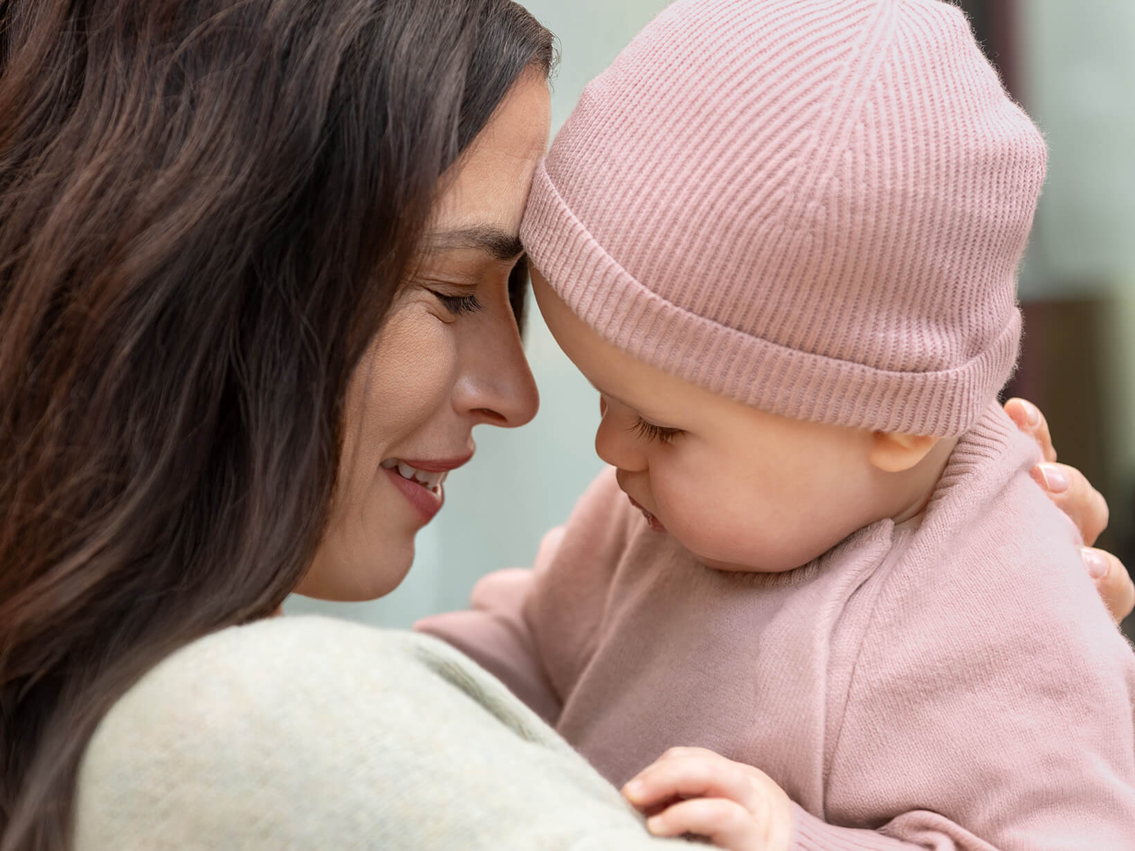 A close up of a woman holding a baby wearing the Nuna Cashmere Two-Piece Sweater Set and Knit Cap in Dusty Mauve Fashion