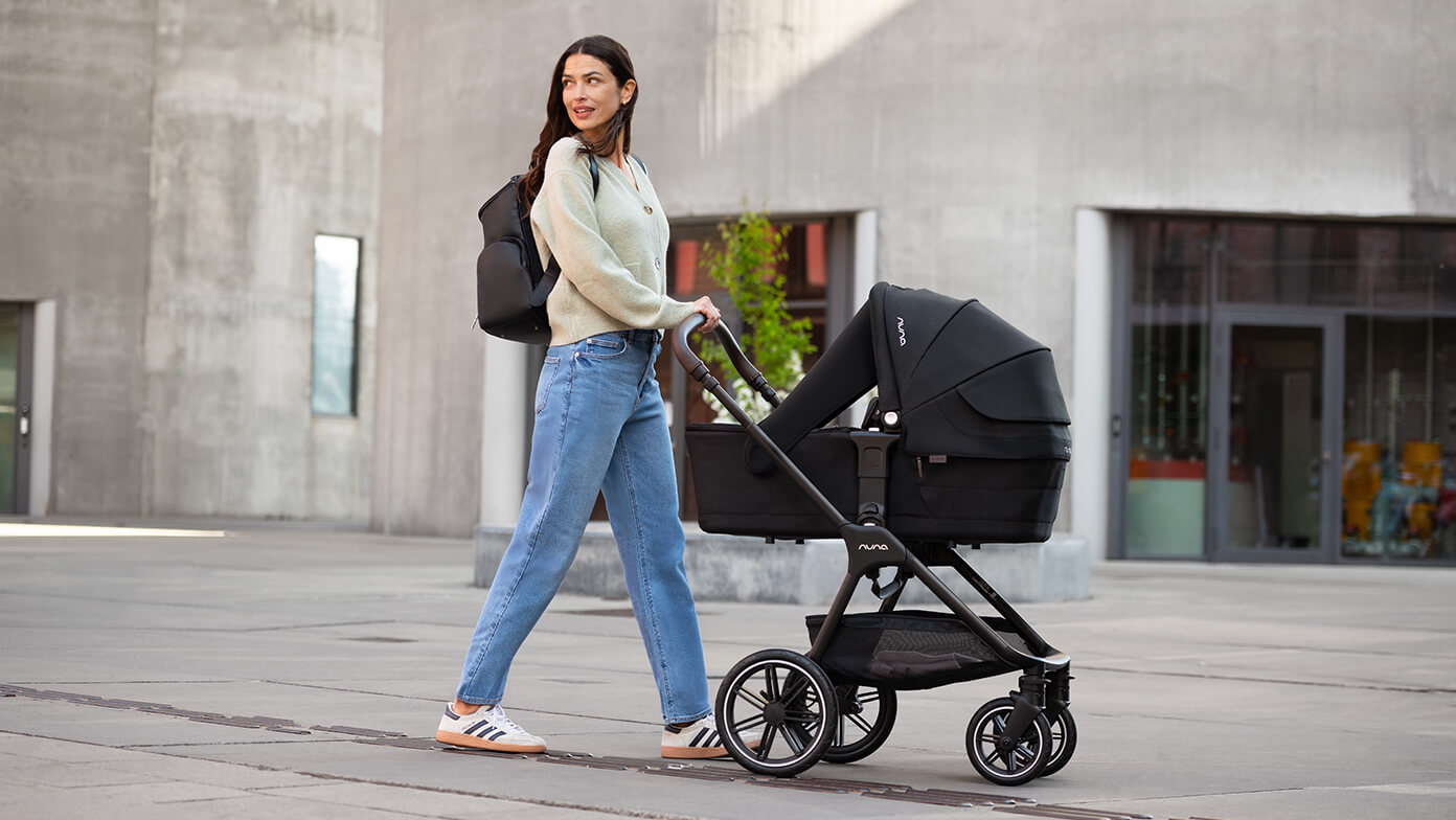 A woman pushes a Nuna pushchair in color caviar with a lie-flat carry cot attached.