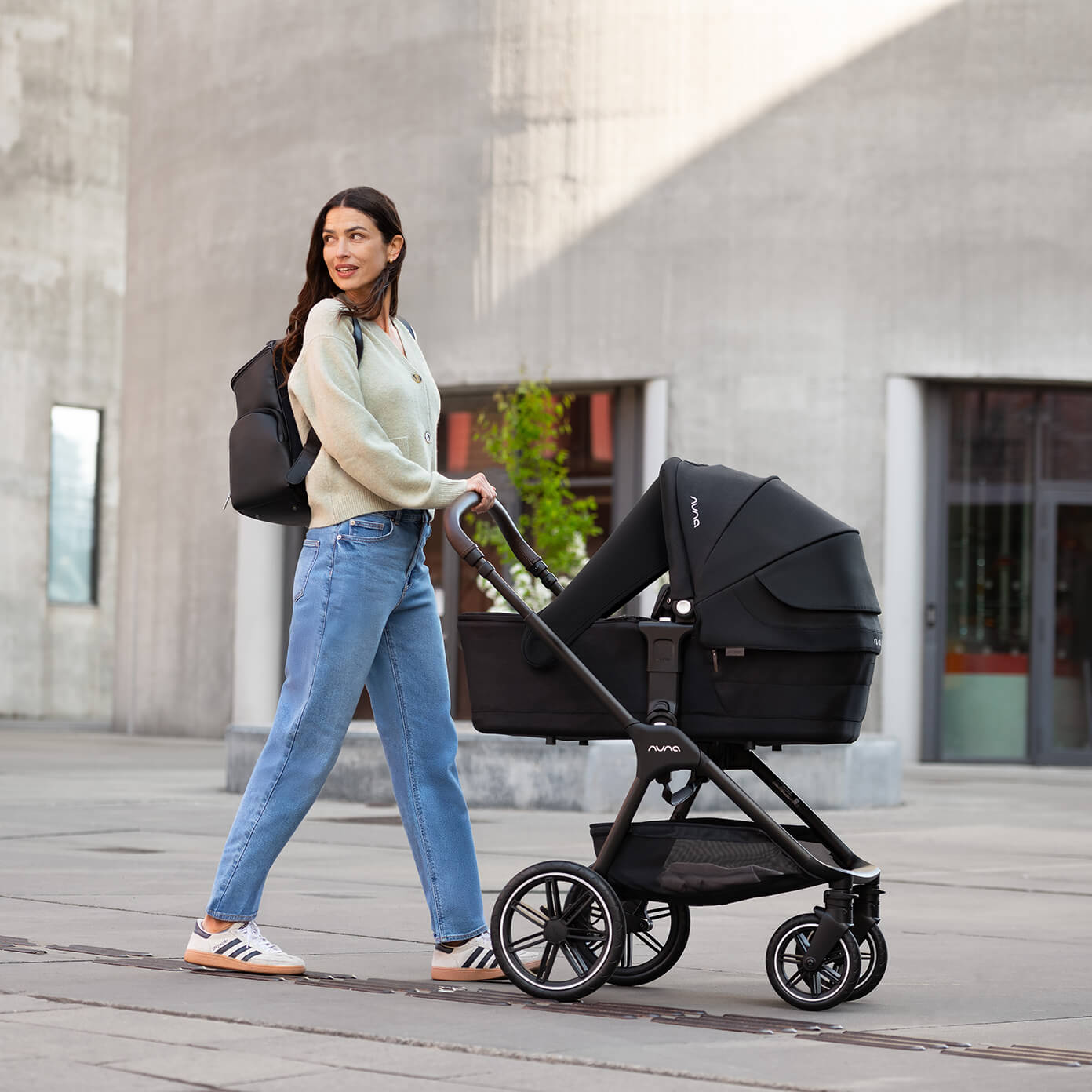A woman pushes a Nuna pushchair in color caviar with a lie-flat carry cot attached.