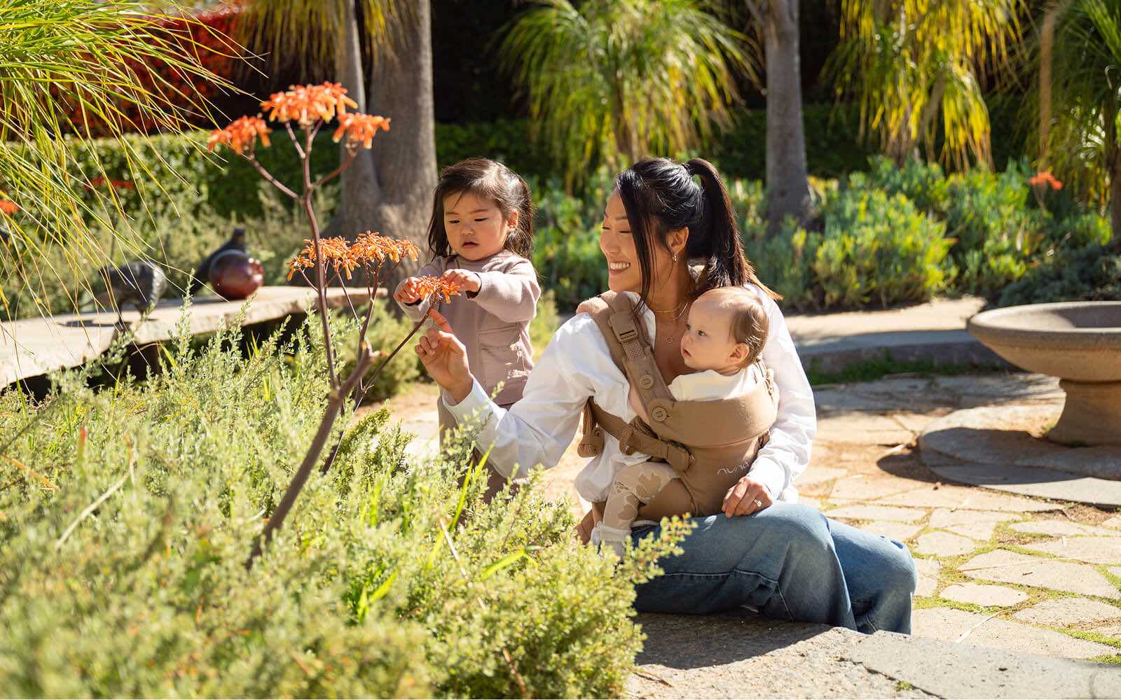 A mother wears a CUDL baby carrier with an infant close to her chest while sitting in a garden beside a toddler.